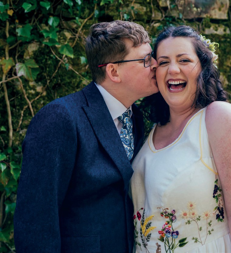 A man in a navy suit kisses a laughing bride on the cheek outdoors, greenery in the background.