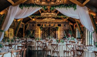 Rustic barn wedding reception with round tables, white linens, wooden chairs, and warm string lights.