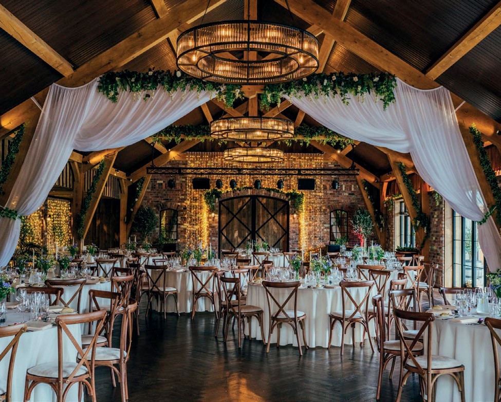 Rustic barn wedding reception with round tables, white linens, wooden chairs, and warm string lights.
