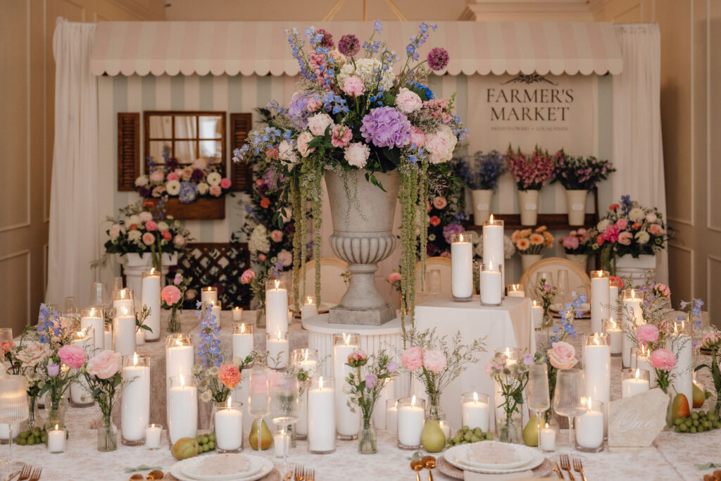 Elegant wedding reception table with a tall floral centerpiece in a gray urn, surrounded by candles and pastel flowers in soft light.