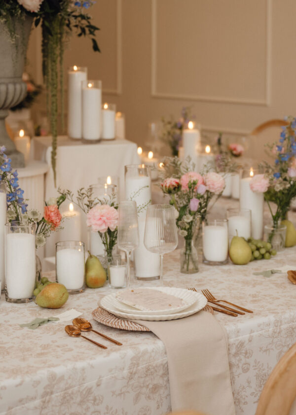 Formal dining table with white candles, glassware, pink and blue flowers, and pears arranged along a lace-patterned tablecloth; gold utensils set at each place.