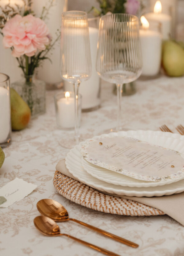 Elegant table setting with stacked white plates on a woven placemat, a floral menu card, and rose-gold utensils, candlelight in the background.