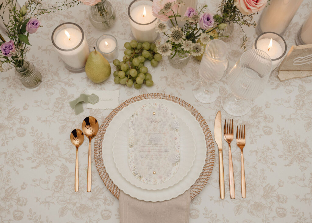 Elegant place setting with white plates, gold flatware, candles, grapes, a pear, and pastel flowers on a patterned tablecloth.