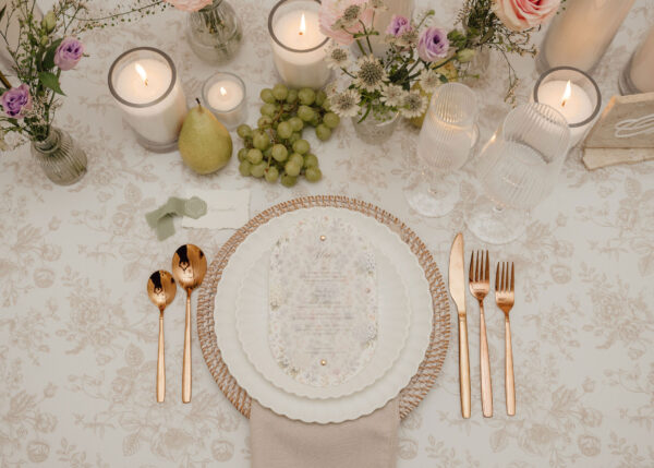Elegant place setting with white plates, gold flatware, candles, grapes, a pear, and pastel flowers on a patterned tablecloth.