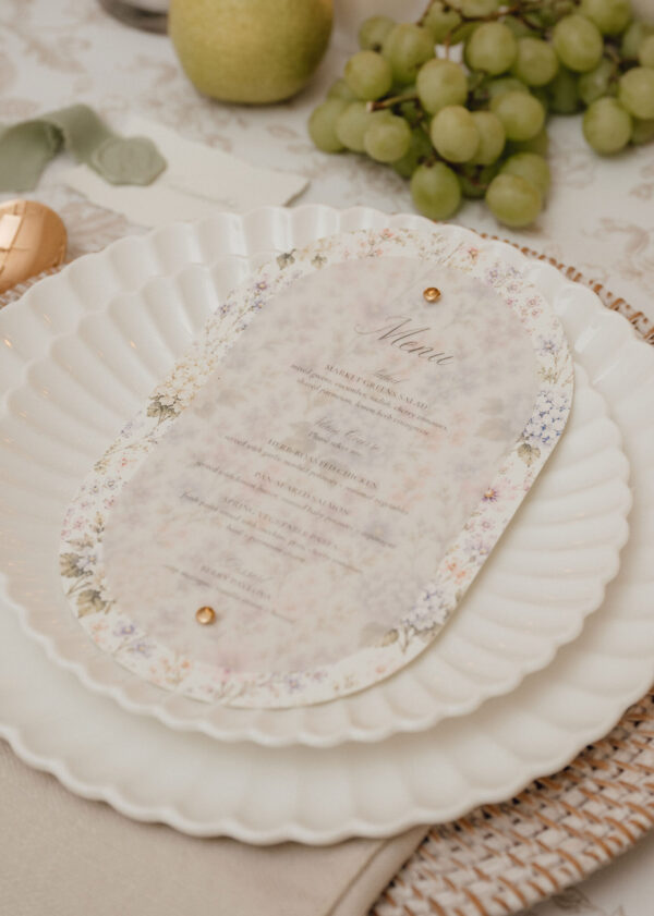 Formal table setting: decorative menu card on a scalloped, floral-bordered plate with a grape cluster in the background.