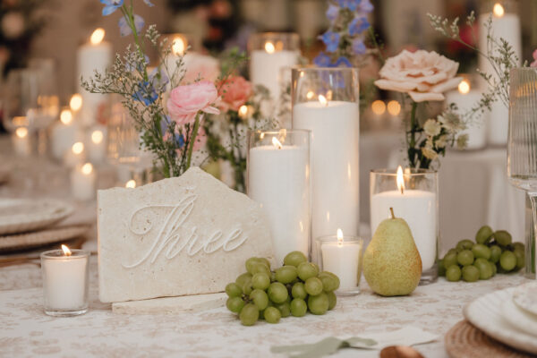Elegant wedding table centerpiece with lit candles, pastel flowers, grape clusters, and a pear centerpiece stone with 'three' engraved.