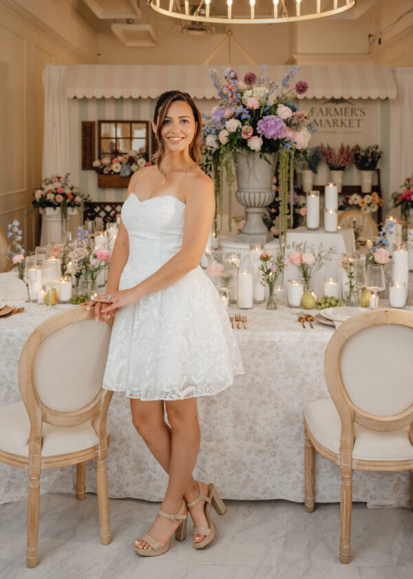 Woman in a white strapless lace dress stands beside a decorated wedding reception table with candles and flowers in a pastel setting.