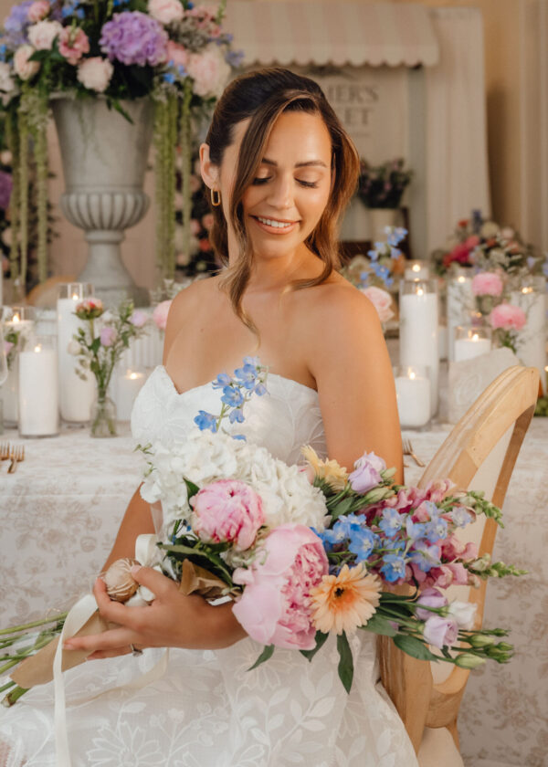 Bride in a white dress sits smiling with a pastel bouquet of flowers in a wedding setting.