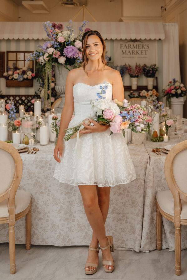 Bride in a white lace dress holding a pastel bouquet, standing at a flower-filled wedding reception with candles and decor behind her, smiling.