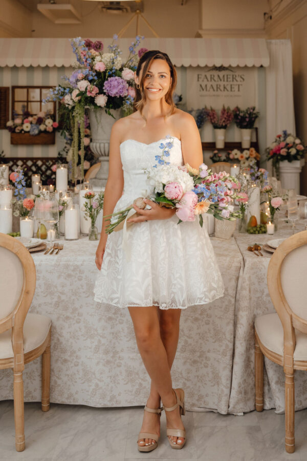 Young woman in a white strapless dress holding a colorful bouquet in a flower-filled banquet hall.