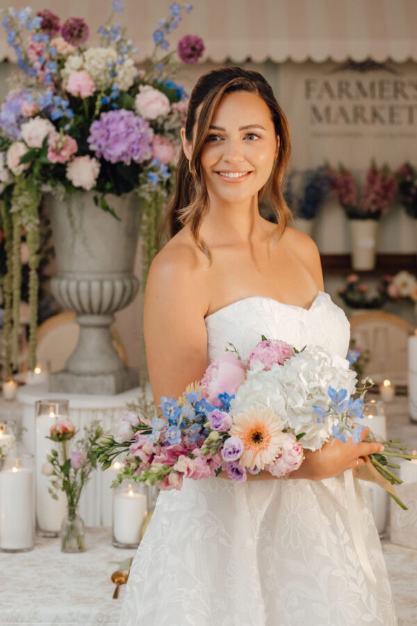 Bride in a white strapless gown holds a pastel bouquet, smiling amid a floral wedding setup with candles in the background.