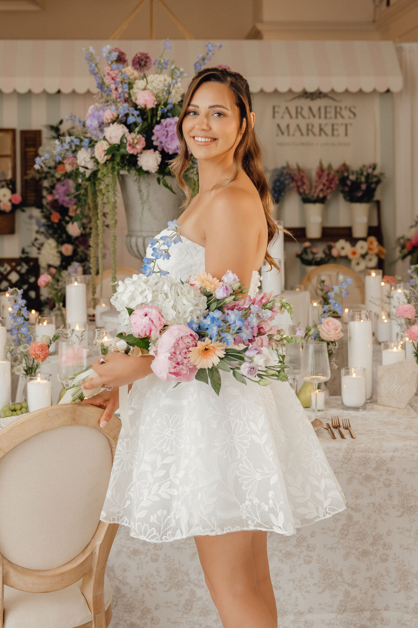 Bride in a white floral dress holding a pastel bouquet at a decorated wedding reception with candles and flowers in the background.