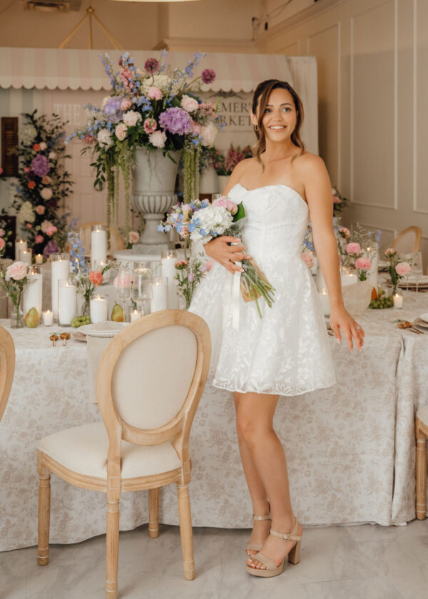 Bride in a white strapless dress holding a pastel bouquet, standing beside a florally decorated table setup in a elegant venue.