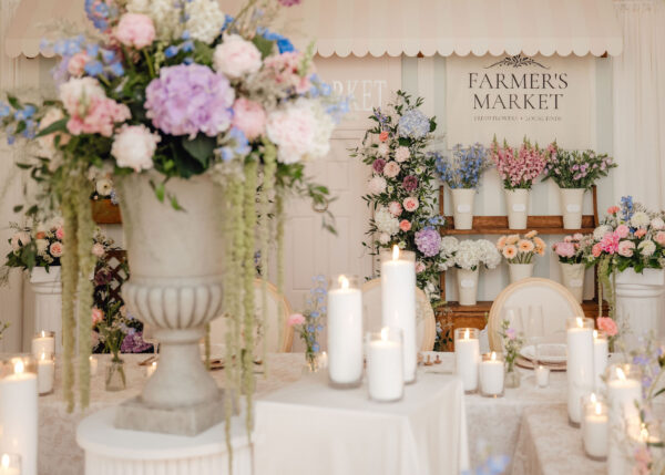 Wedding reception decor with abundant pastel flowers, white candles, and a 'Farmer's Market' sign in the background.
