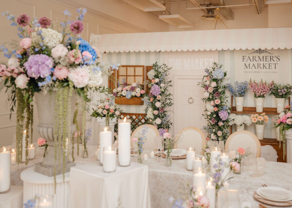 Elegant pastel floral market display with white candles on lace-covered tables and a 'Farmer's Market' backdrop.