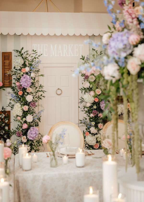 Pastel wedding or event table setup with tall floral arrangements and candles, soft lace tablecloth, and a pale door marked 'THE MARKET' in the background.