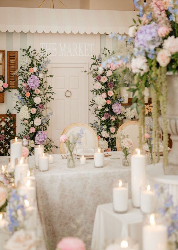 Pastel floral wedding decor with tall arrangements, candles on a lace-covered table, and a pale doorway in the background.