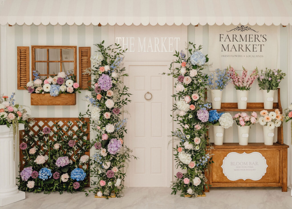Pastel storefront with striped walls, white door, and abundant pink, blue, and white flowers in wooden planters and arrangements.