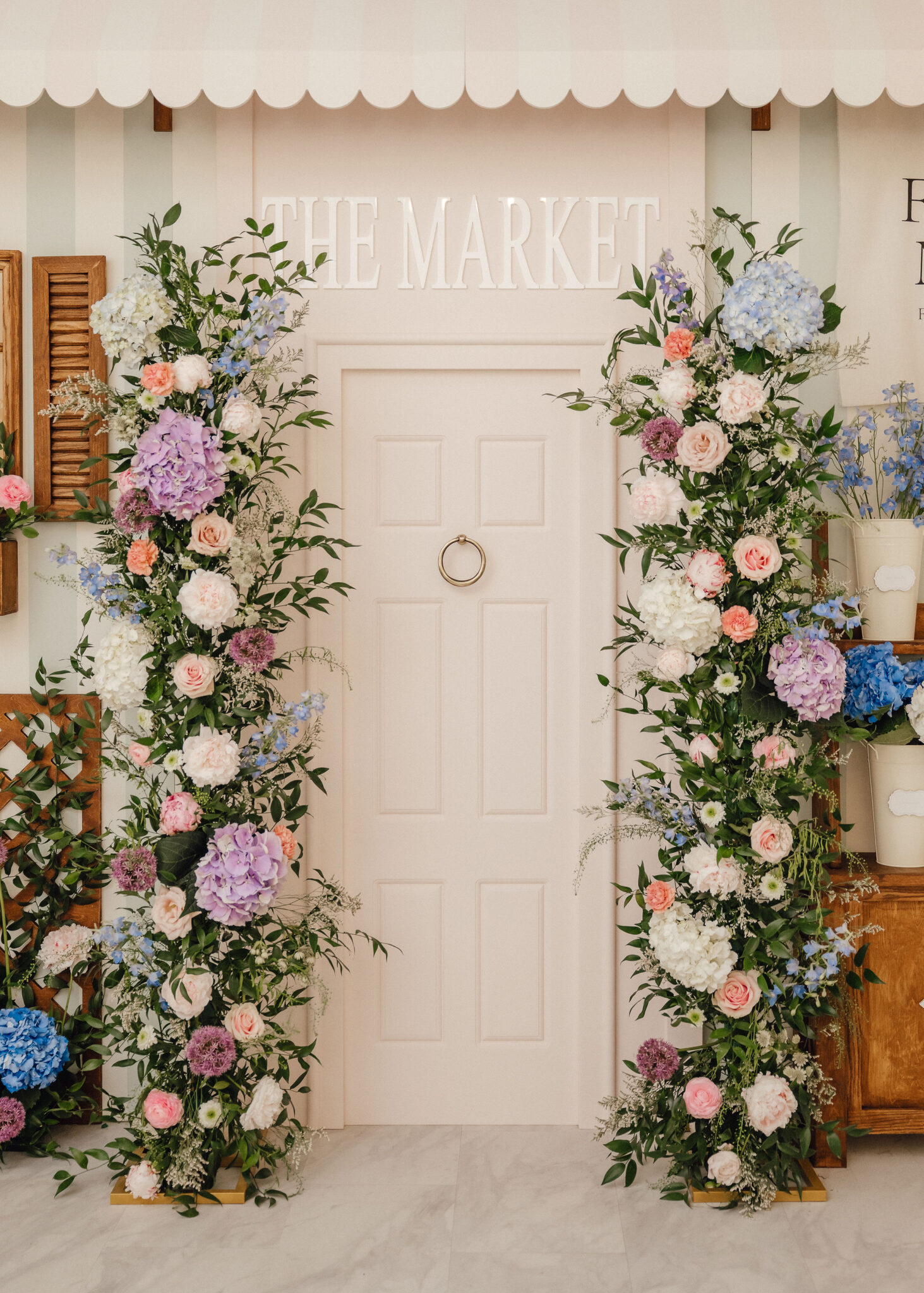 Pastel floral arrangement framing a white door with a sign reading 'THE MARKET' above it, creating a decorative entrance.