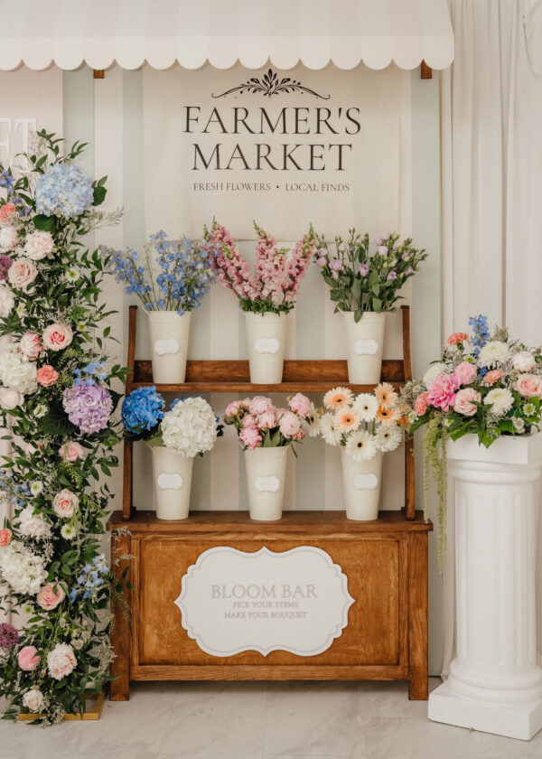 Flower display at a market stand: pastel bouquets in cream buckets on a wooden shelf under a striped canopy with a 'Farmer's Market' sign above (Fresh Flowers, Local Finds).