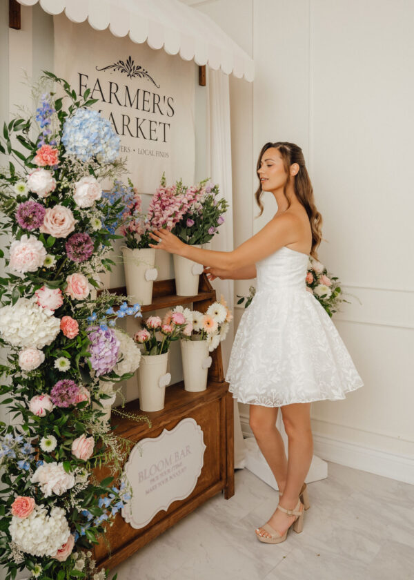 Bride in a white dress selects pastel flowers from a large floral display beneath a 'Farmer's Market' sign framed by a scalloped canopy.