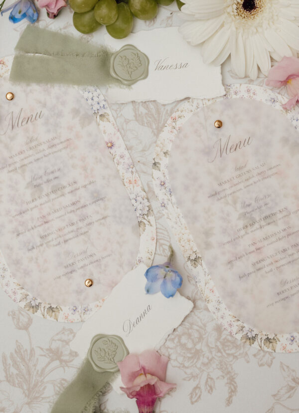 Flat lay of a vintage wedding invitation suite with wax seals, torn name tag reading Vanessa, floral borders, and pastel flowers and ribbons.