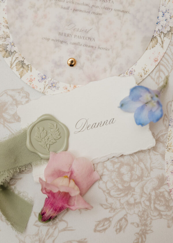 Close-up of a vintage wedding invitation with floral edges, a gold wax seal, and the name Deanna in script