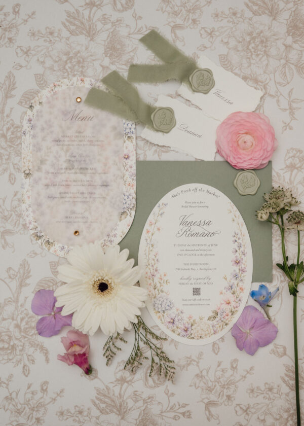 Elegant wedding invitation suite with pale green wax seals, torn-edge cards, and muted floral backdrop on a patterned tablecloth.