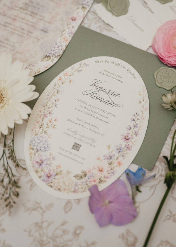 Round wedding invitation with delicate floral border and script names Vanessa Romando on a pale background, surrounded by flowers and greens.