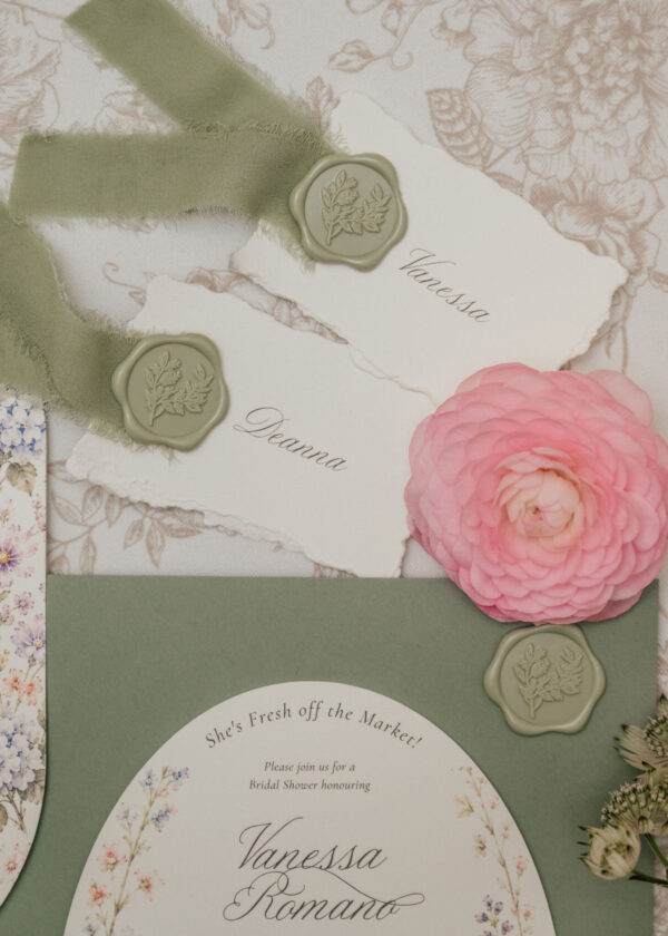 Soft pastel wedding invitation with sage-green ribbon, wax seals, and pink ranunculus on a floral backdrop