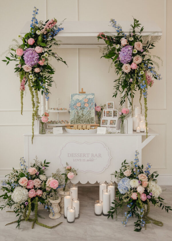 Floral arch decorations surrounding a white dessert display with pastel flowers and candles for a wedding or event setup.