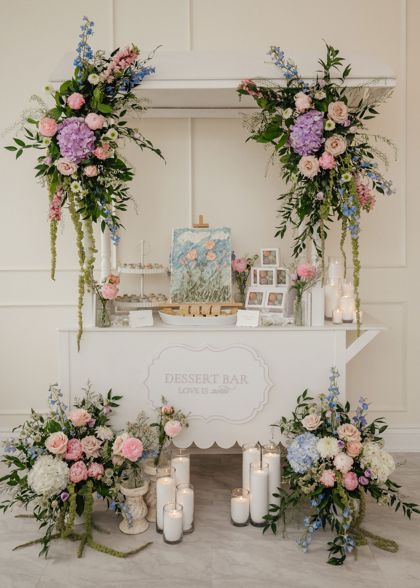 Floral arch decorations surrounding a white dessert display with pastel flowers and candles for a wedding or event setup.