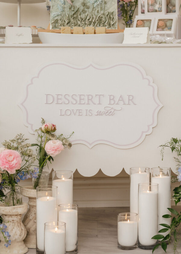 White dessert bar with a pastel-pink framed sign reading 'DESSERT BAR', flanked by pink roses, greenery, and tall candles in glass holders