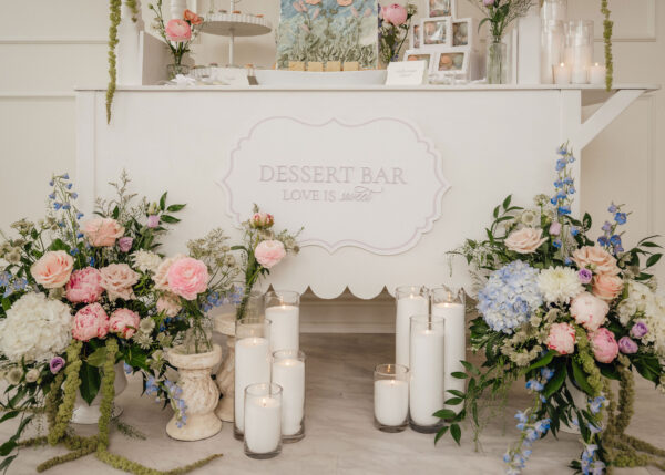 Elegant dessert bar adorned with pastel flowers and tall white candles on a white tasting table.