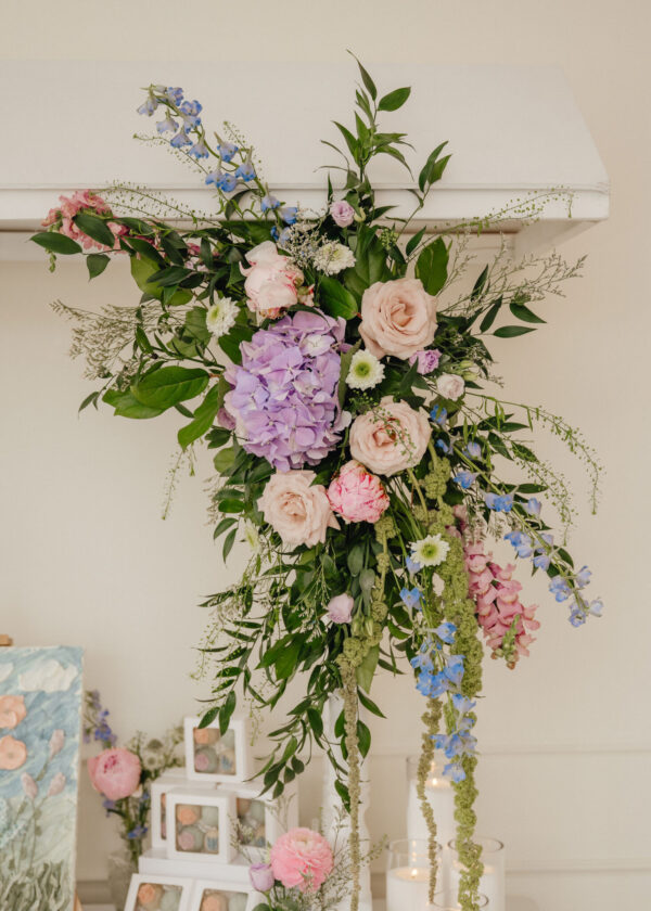 Hanging floral arrangement with pastel roses, hydrangeas, blue delphiniums and greenery cascading against a pale wall.