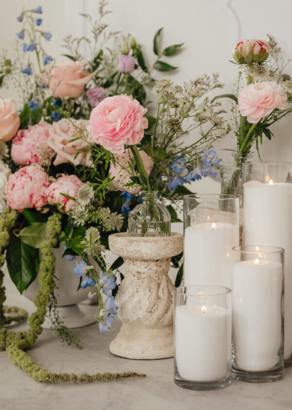 Pastel floral centerpiece with pink peonies and blue accents in glass vases beside white candles on a stone pedestal table.