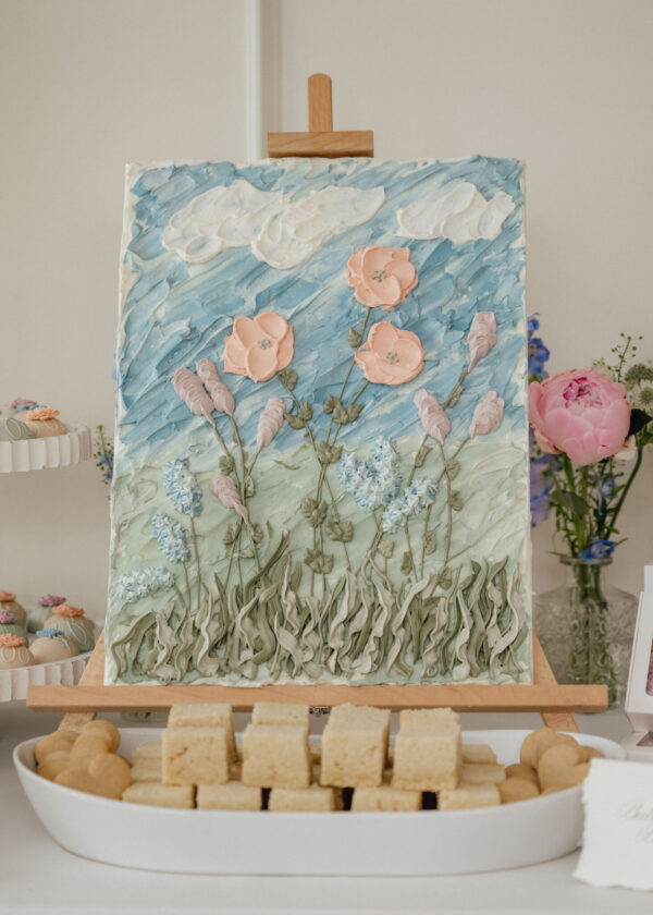 Large decorative cake on a wooden pedestal with blue frosting, pink sugar flowers, and greenery, flanked by pastries on the table