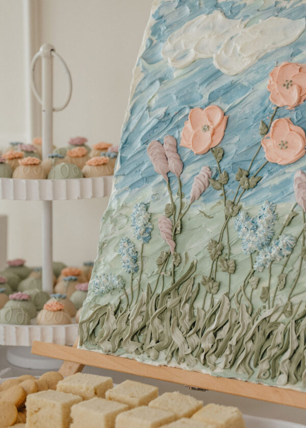 Close-up of a large multi-layer cake with blue-green icing and pink sugar flowers, in a bakery display with cupcakes visible in the background.