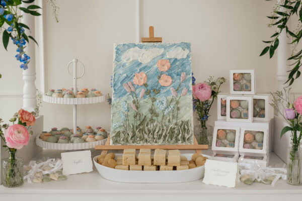 Pastel dessert table with a floral textured cake on a wooden stand, surrounded by cupcakes, pastries, and pink roses in a bright setting.