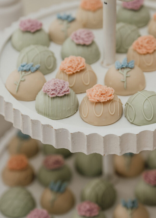 Pastel fondant cupcakes with flower toppers in green, peach, and pink, arranged on a white display tray by colorful rosettes.