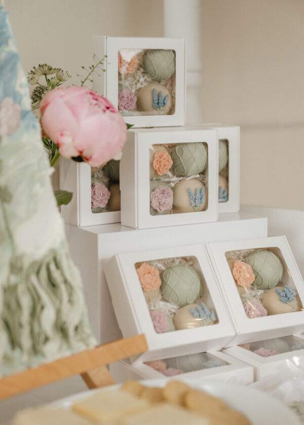 Boxes of pastel cookies with floral decorations in clear windows, arranged on a white display shelf in a soft setting.