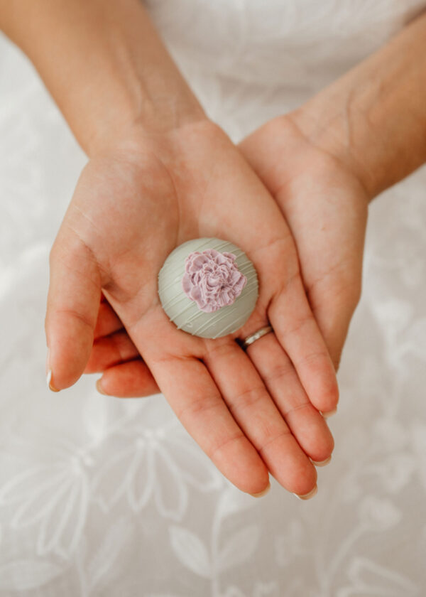 Hands cupping a round gray fondant-covered treat with a pink rose on top, held over a white background