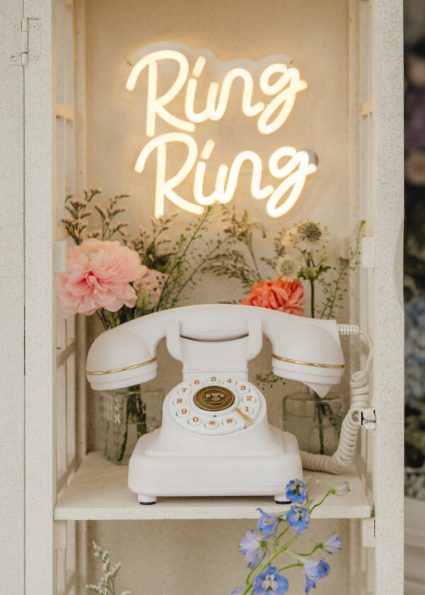 White rotary dial telephone in a display cabinet with a glowing 'Ring Ring' neon sign and pastel flowers nearby.