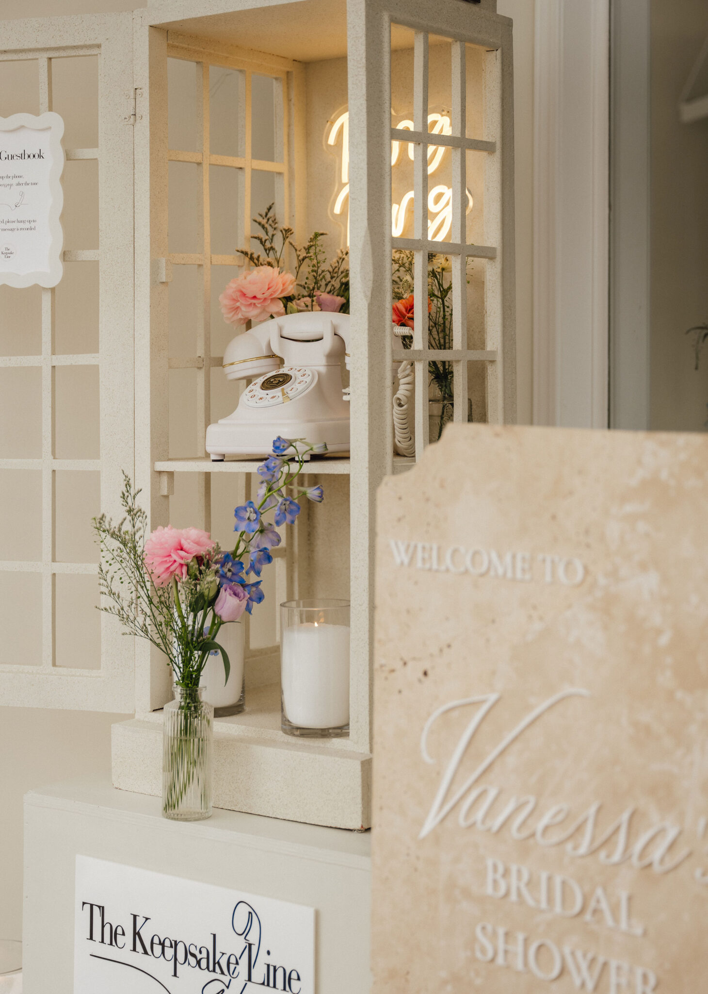 Beige lattice display with a vintage white rotary phone, glass vases of pink and blue flowers, and a warm neon sign in the background—creating a bridal shop display.