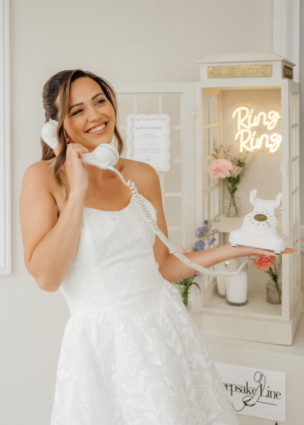 Smiling bride in a white lace dress holds a vintage white telephone, standing beside a pastel bridal shop display with a glowing 'Ring Ring' sign and flowers behind her.