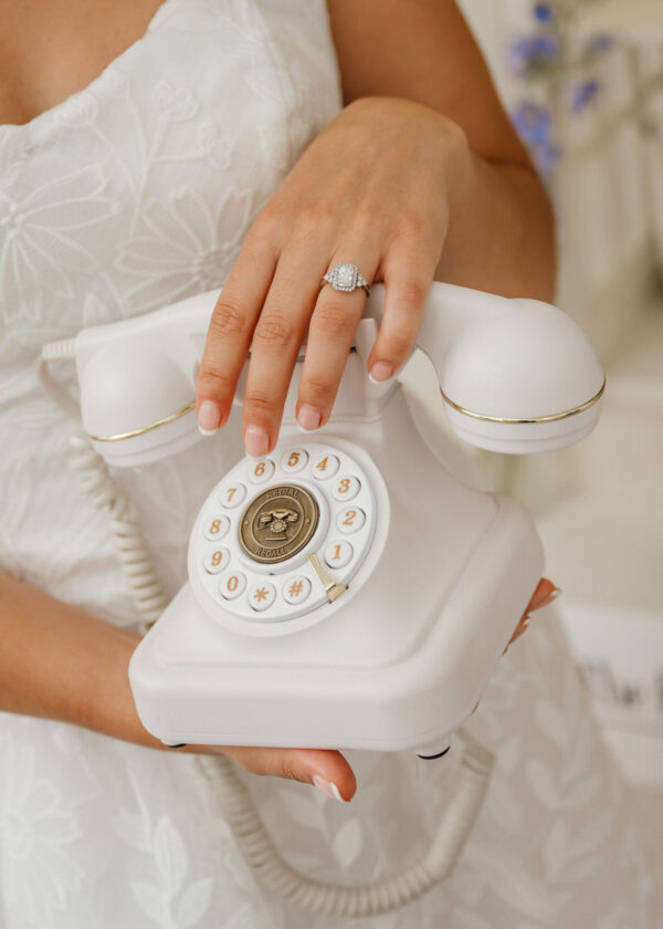 Bride's hand resting on a white rotary telephone, showcasing a diamond engagement ring against a lace wedding dress.