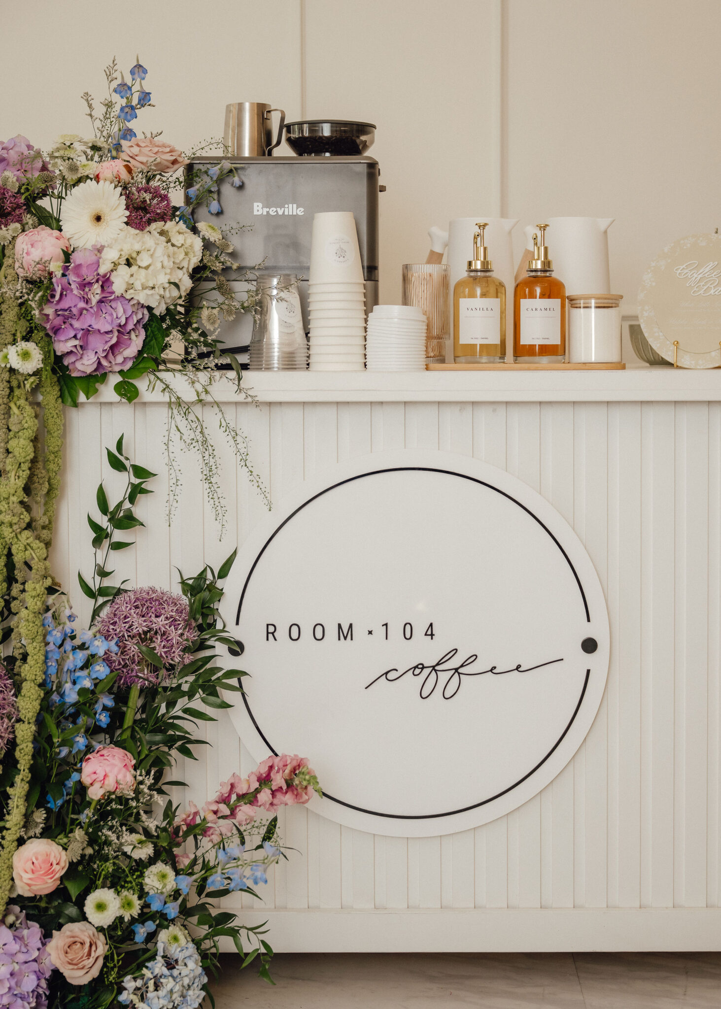 Floral arrangement beside a Breville coffee machine and stacked cups on a white counter, with a large round ROOM 104 coffee sign nearby