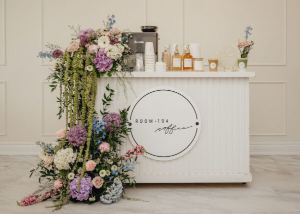 Colorful flower arrangement drapes over a white coffee bar with a Breville espresso machine in a neutral, sunlit room.