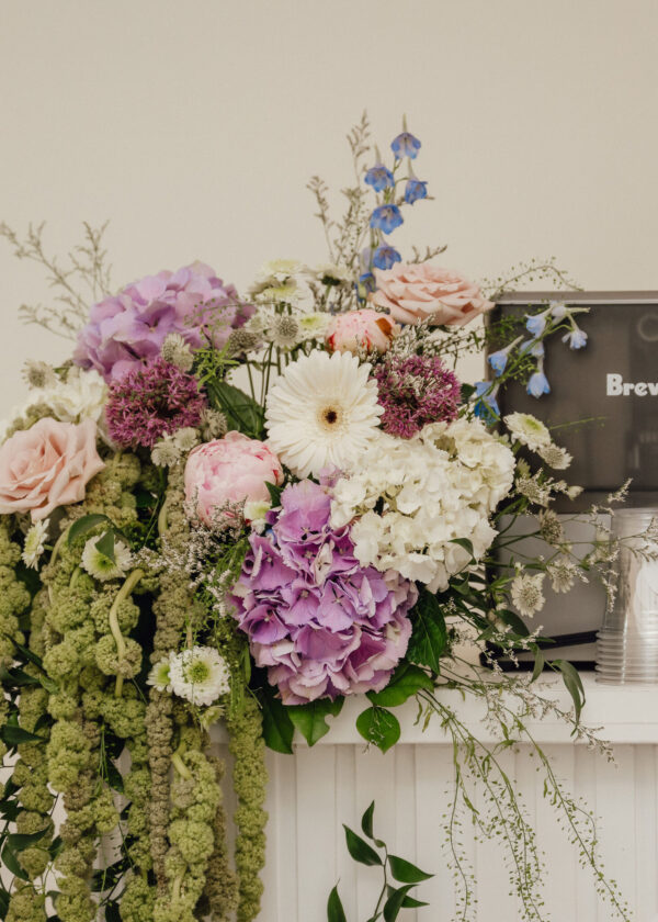 Colorful bouquet with pink roses, purple hydrangeas, white gerbera, blue delphiniums, and cascading greenery against a pale background.