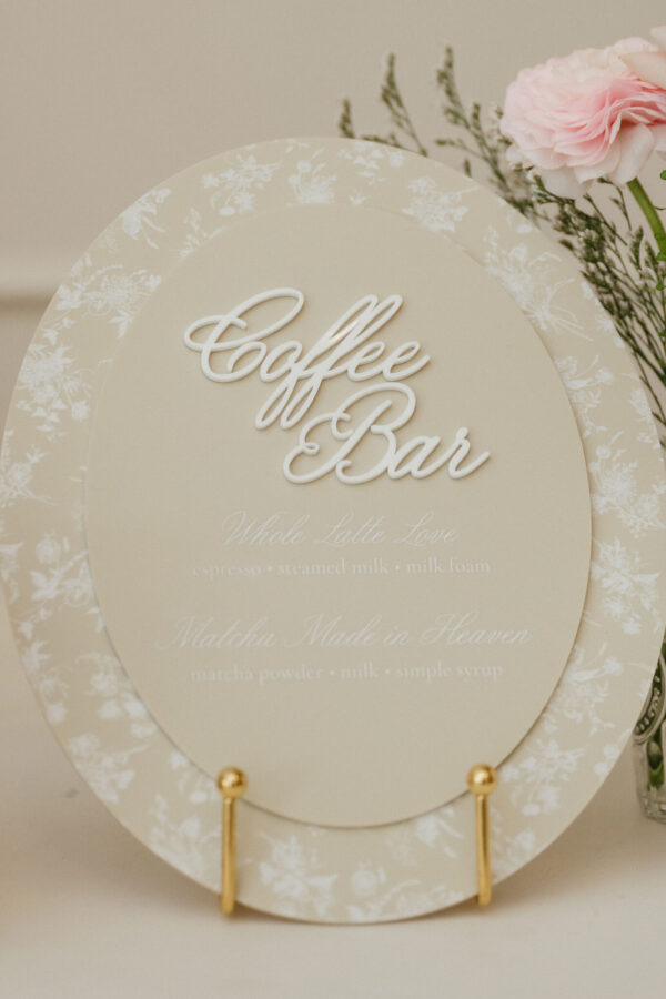 Beige circular sign with white cursive text 'Coffee Bar' and a floral edge, accompanied by pale pink flowers nearby (decorative display).
